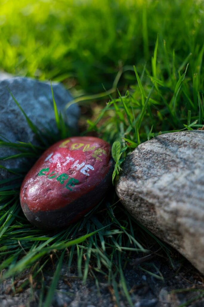 Colorful painted rock with 'Love is the cure' lies in lush grass surrounded by stones.